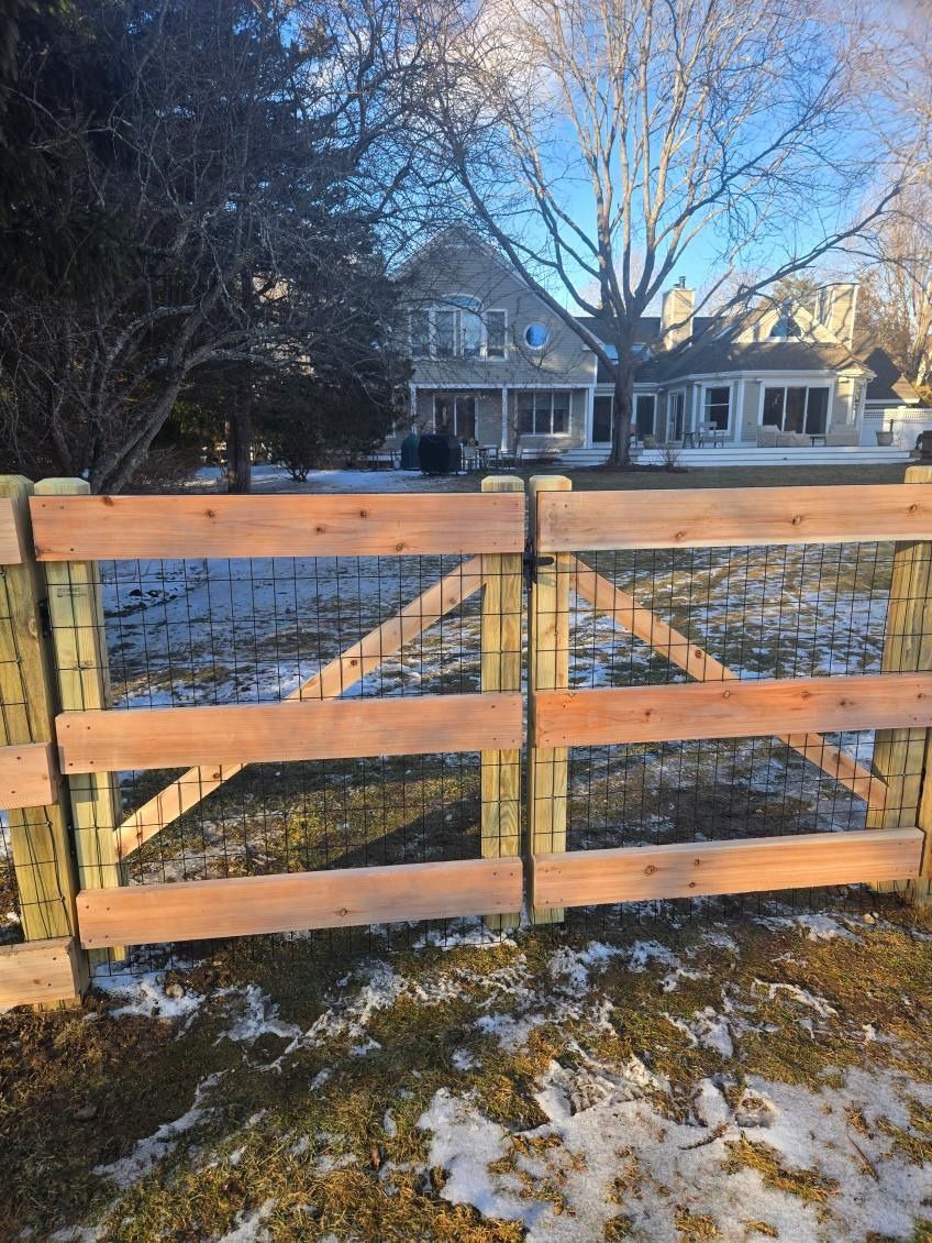 Wooden farm gate in a snowy yard, with a house and leafless trees in the background.