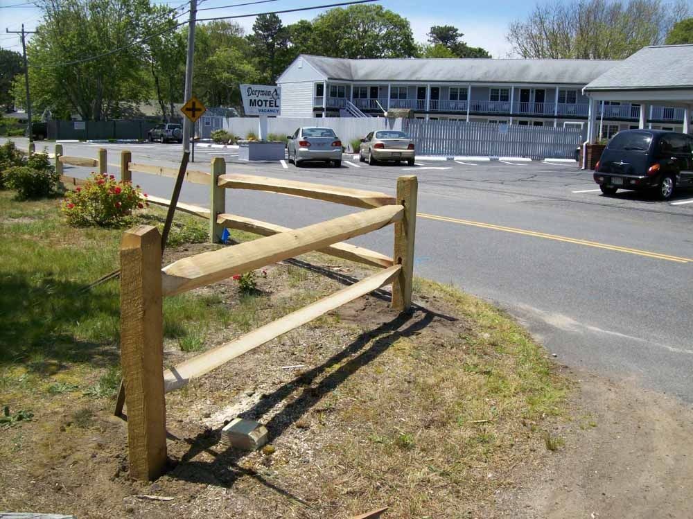 Wooden roadside barrier beside a two-lane street near a white building and parked cars
