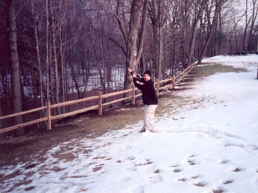 Person in dark jacket standing on a snowy path beside a wooden fence in a leafless wooded area