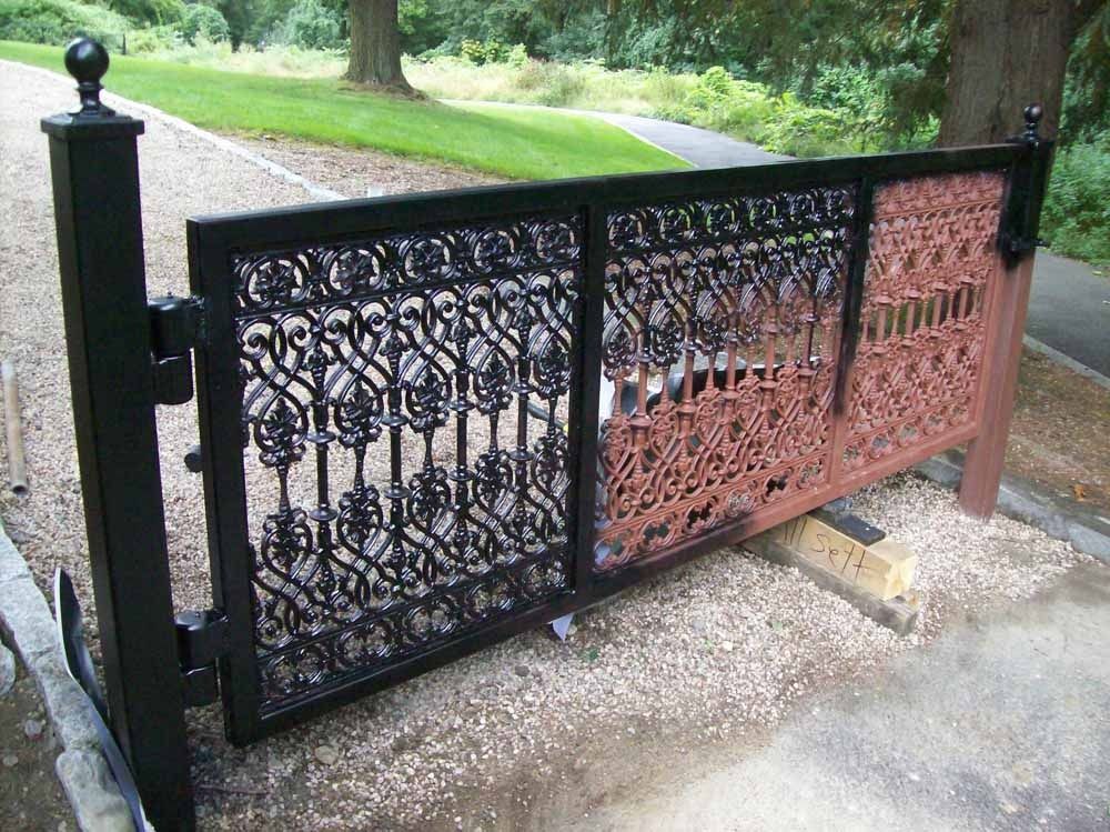 Decorative black metal gate with a reddish-brown panel beside a gravel driveway and green lawn