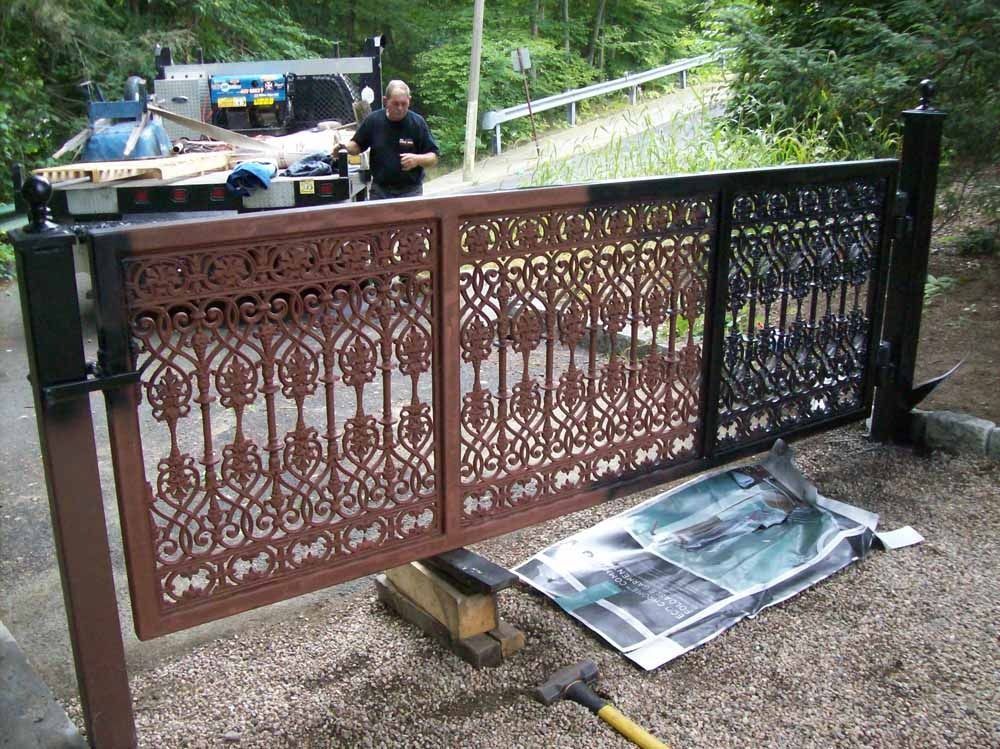 Damaged ornate metal gate lying partly detached on a wet path, with a vehicle and two people in the background