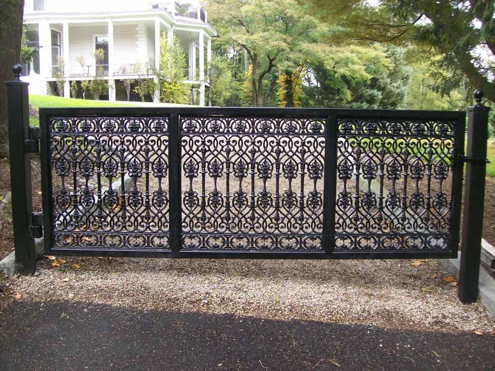 Black ornate metal driveway gate in front of a house and trees