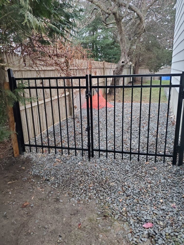 Black metal gate blocking a gravel driveway beside trees and a house