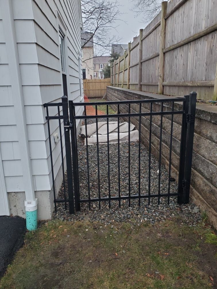 Narrow gravel side yard with a black metal gate between a house and wooden fence.