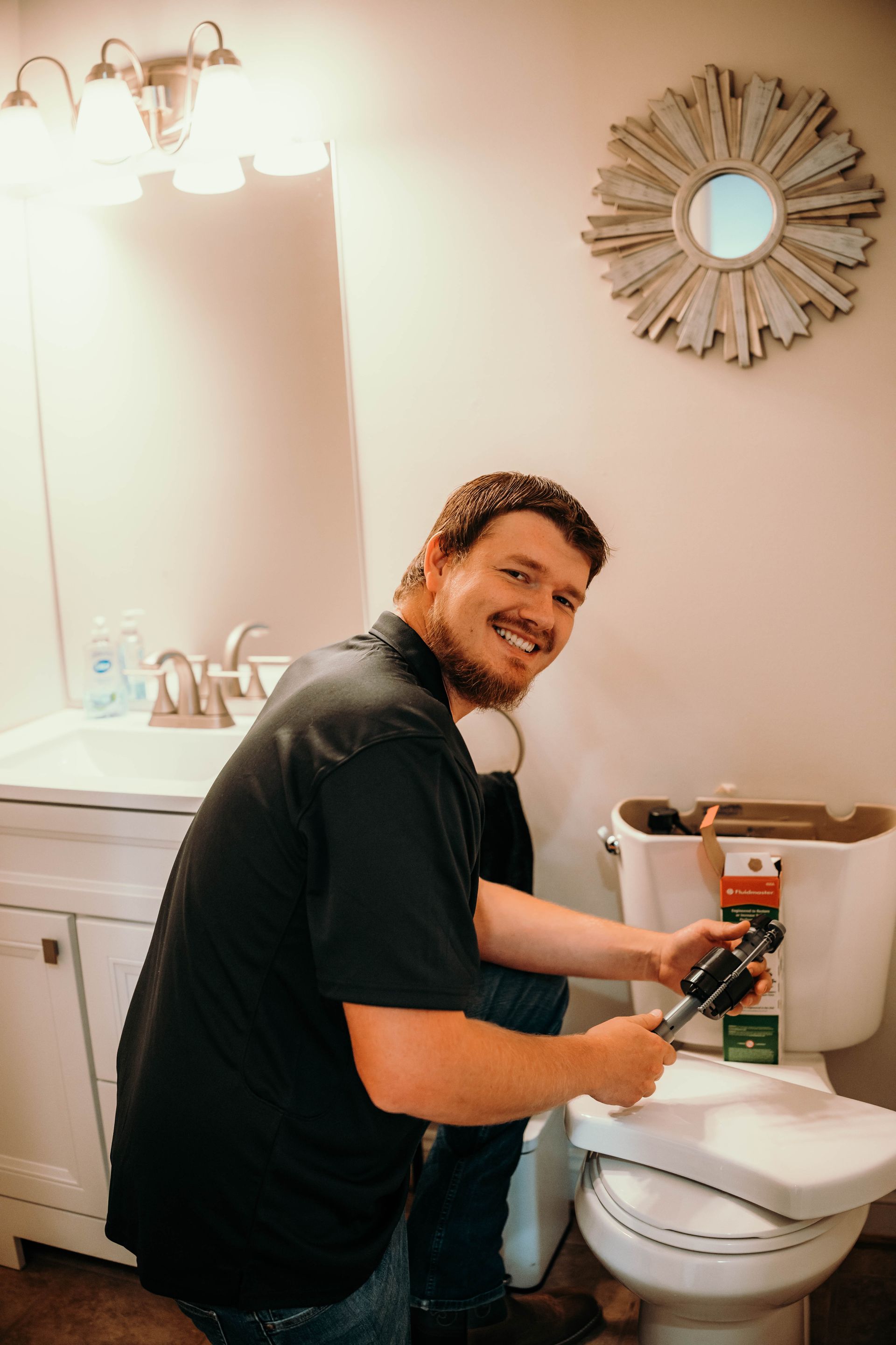 Man fixing a toilet in a bathroom, smiling. Holds tool, wearing black shirt.