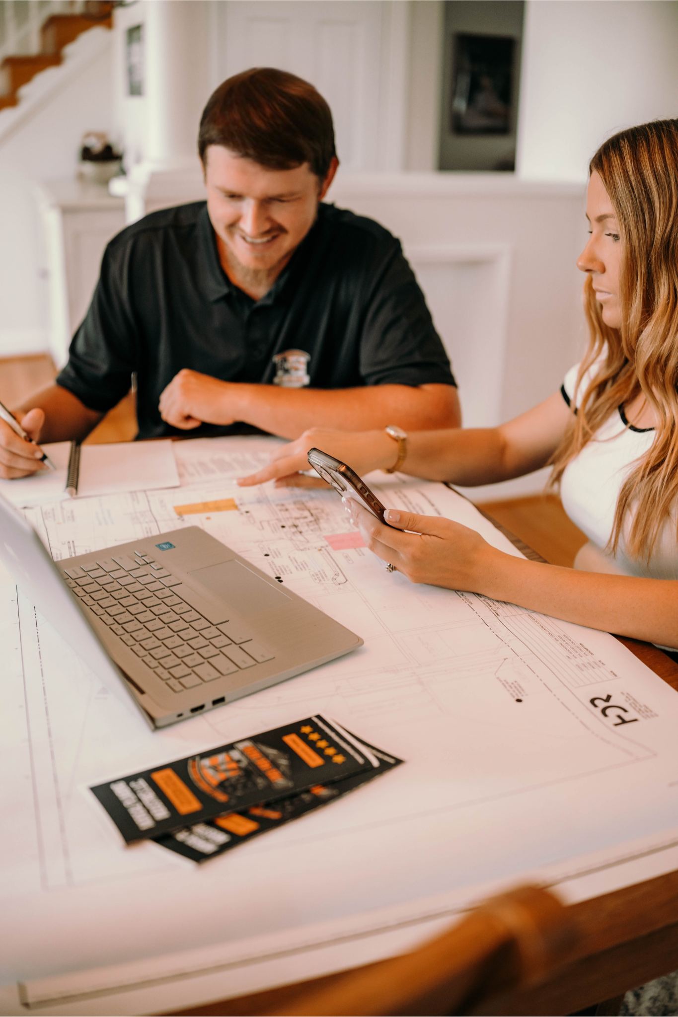Two people reviewing blueprints at a table with a laptop and brochures.
