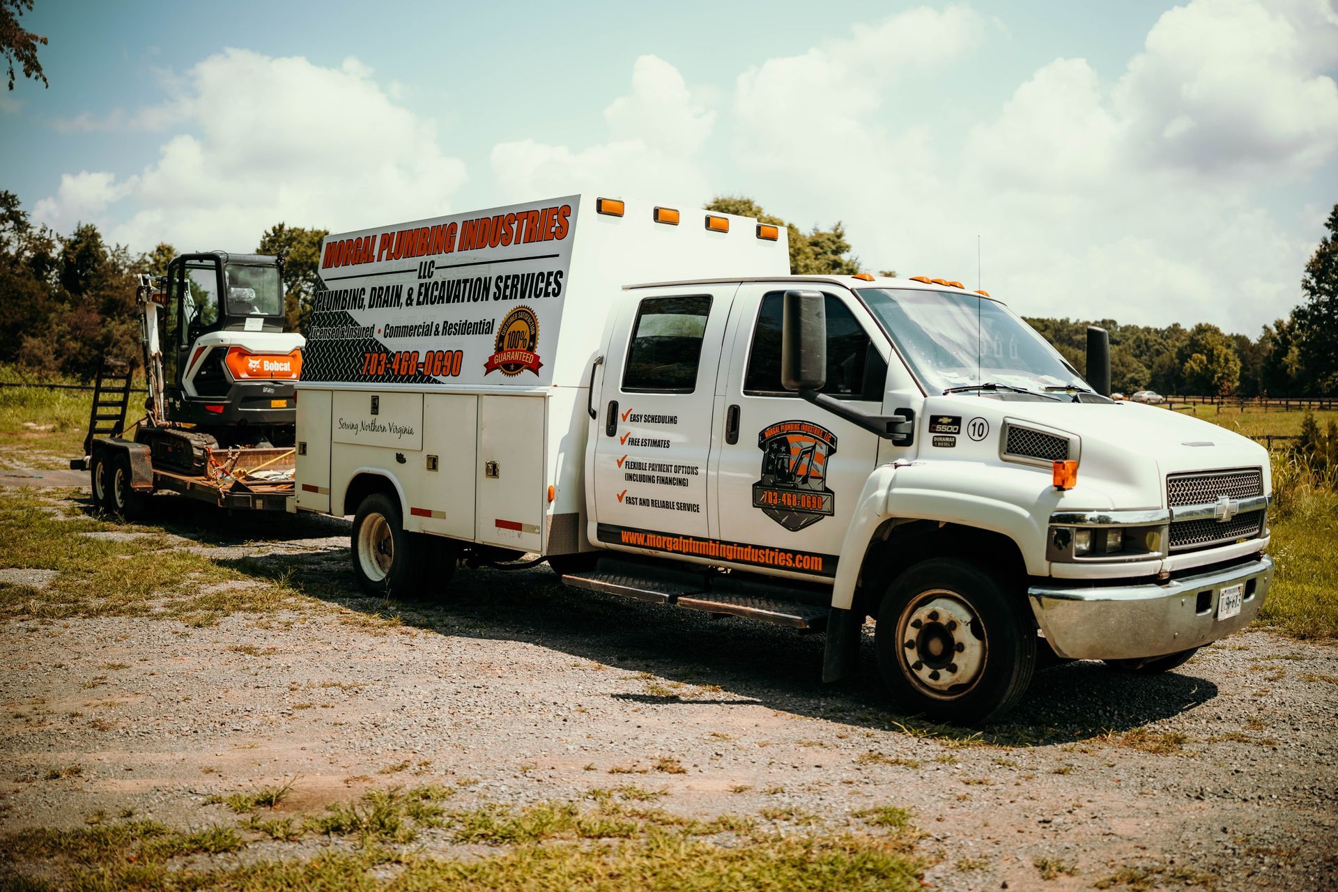 White service truck with trailer carrying excavator parked outdoors on gravel.