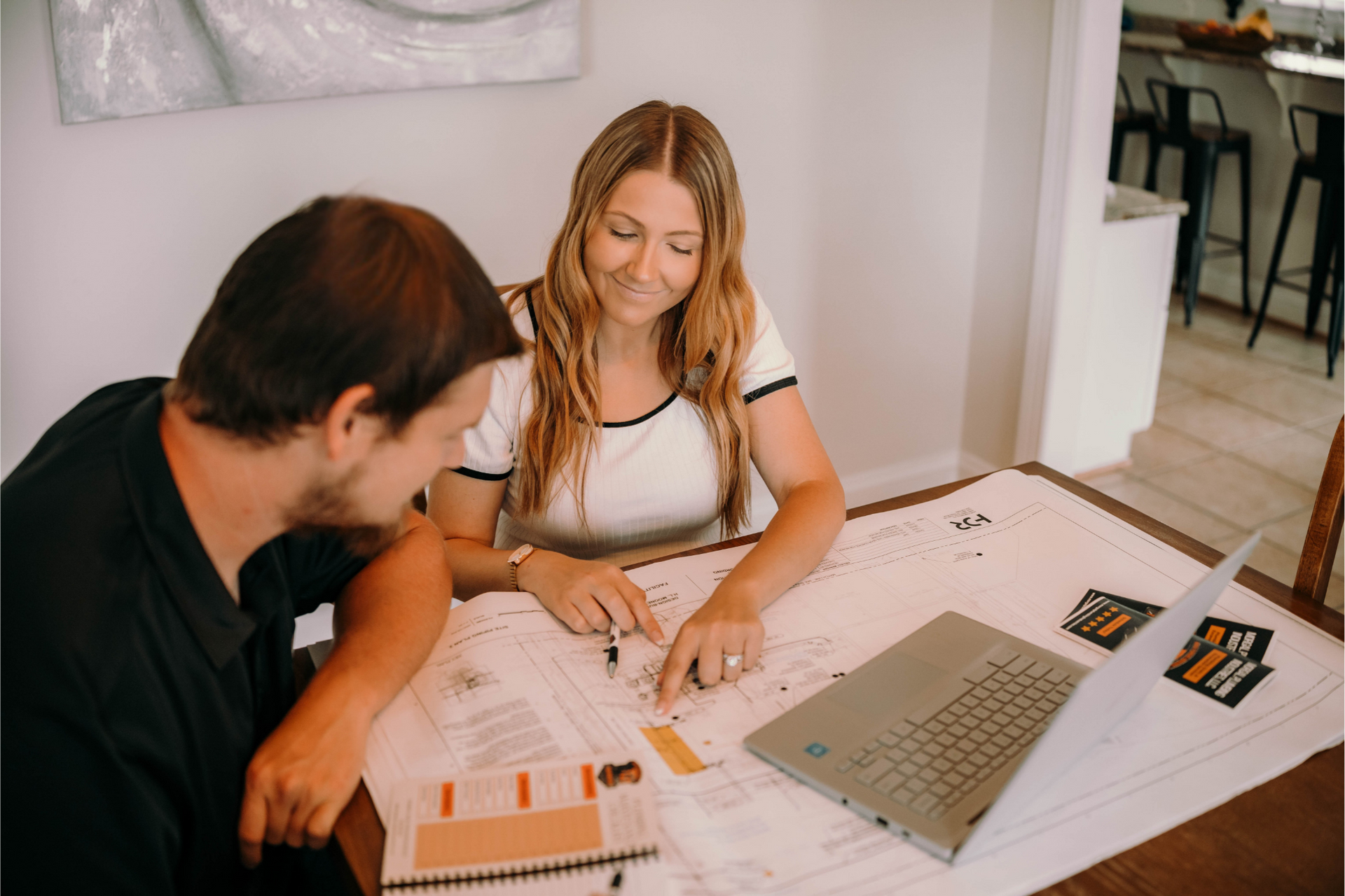 Couple reviewing blueprints at a table with a laptop, smiling and pointing.