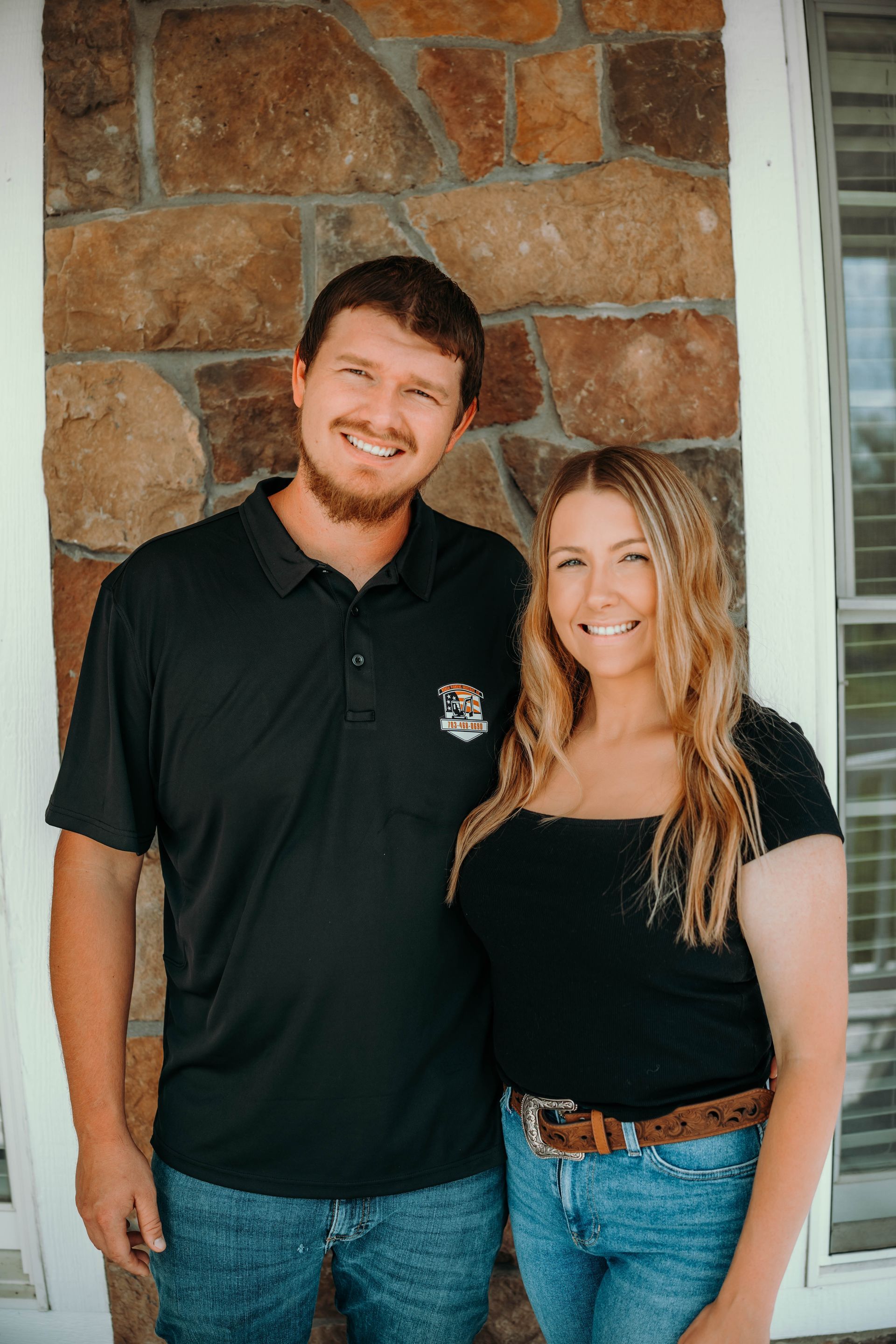 A man and woman smile, standing together near a stone wall and window.