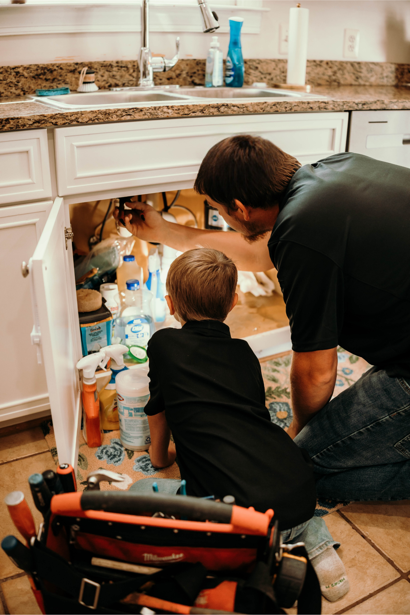 Man and child working under a kitchen sink, examining pipes. A toolbox rests on the floor.