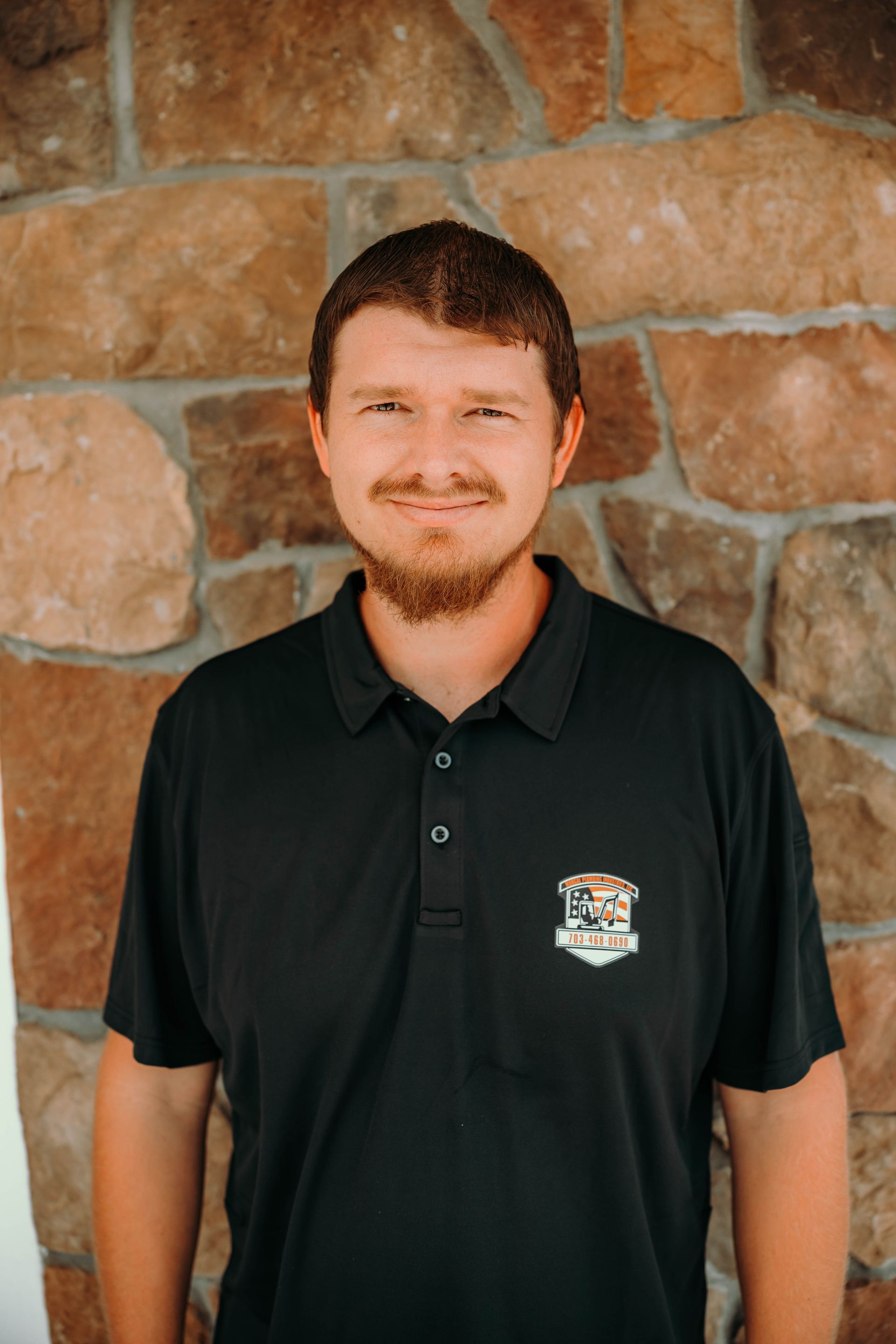 Man with a beard smiles in a black polo shirt against a stone wall.