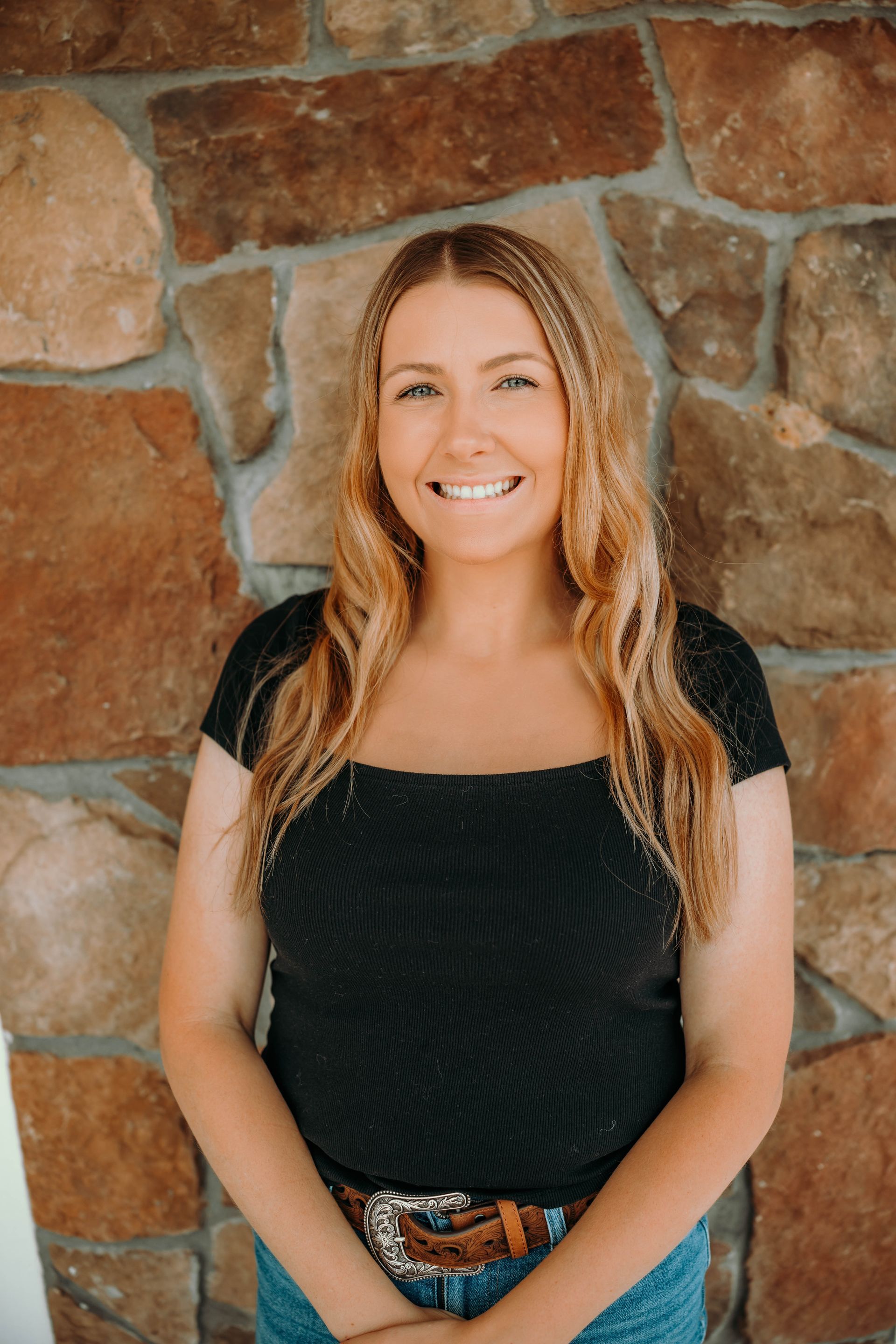Woman with long blonde braids smiles in front of a stone wall, wearing a black top and jeans.