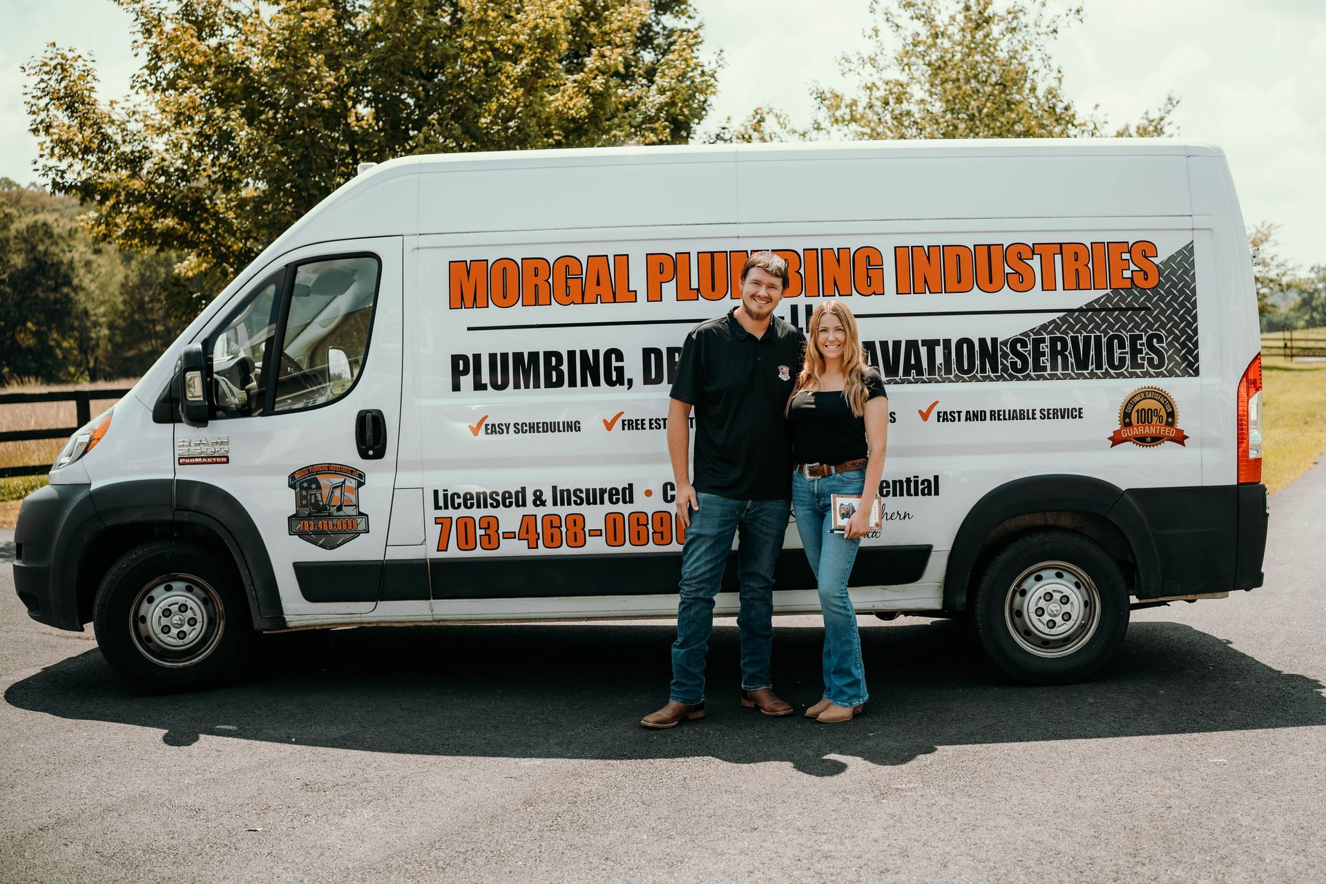 A man and woman stand next to a white work van with business branding. They are on a paved road.