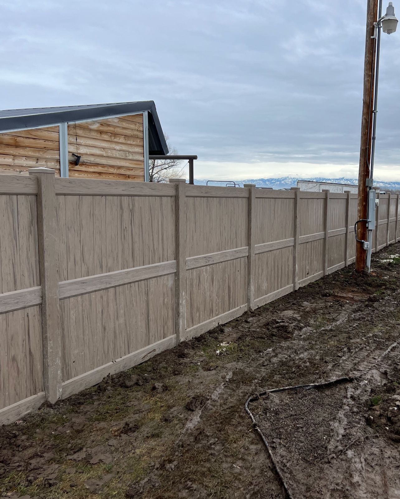 Tan vinyl fence in front of a building and power pole against a cloudy sky.