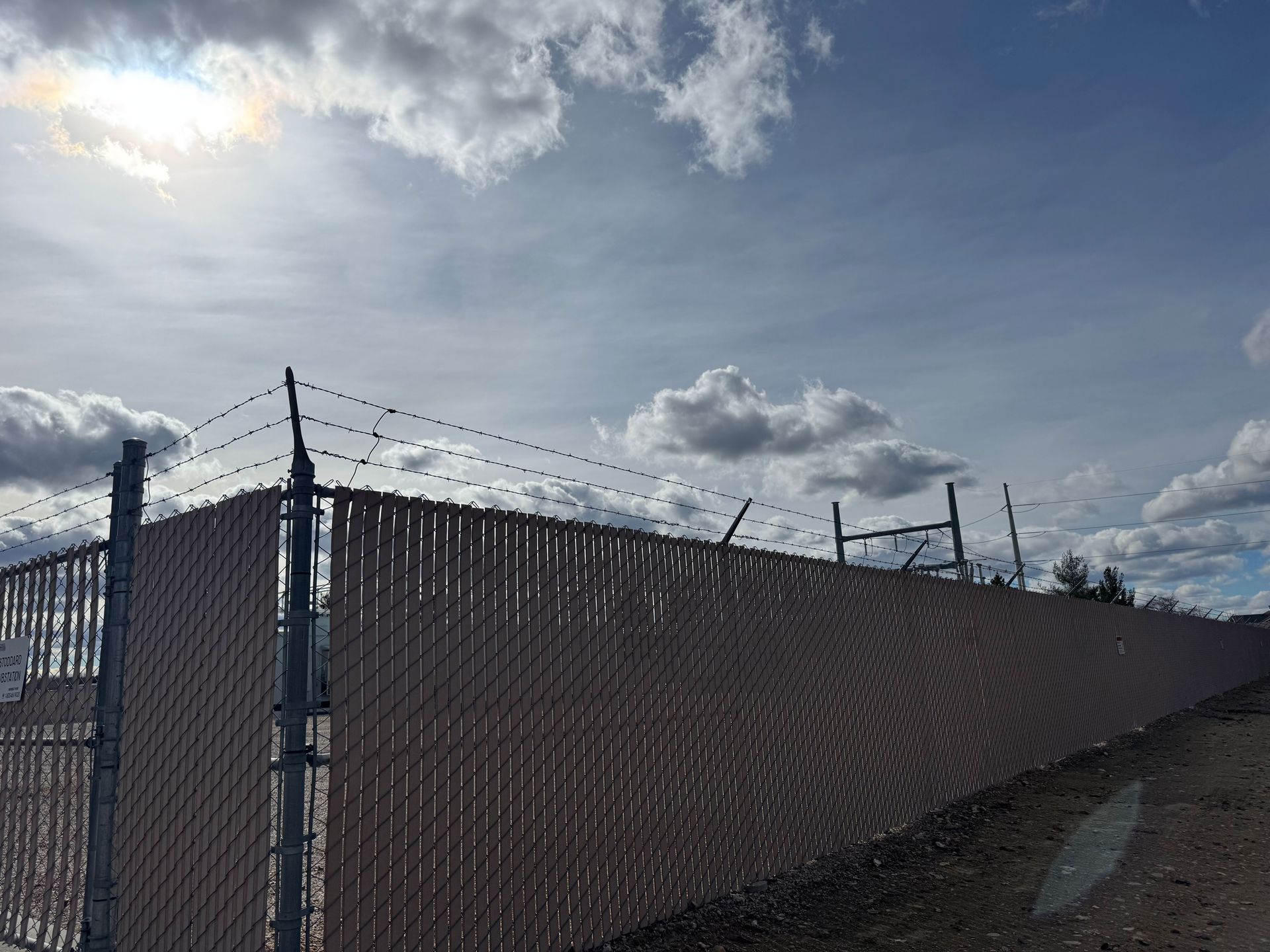 Chain-link fence with brown privacy slats and barbed wire under a cloudy sky.