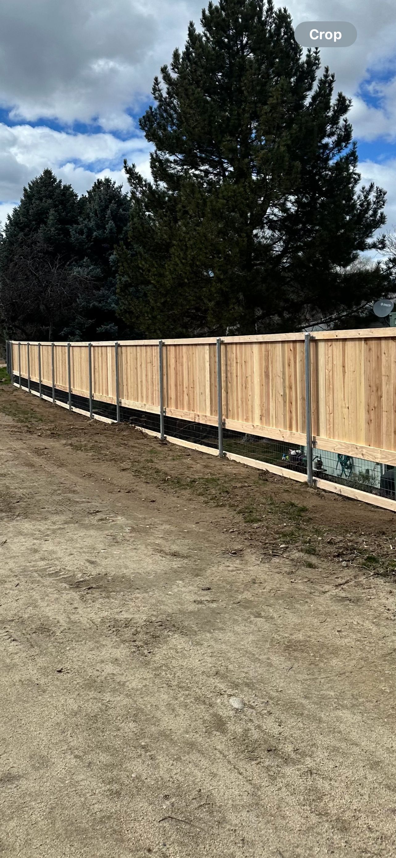 Wooden fence along a gravel area with trees and a cloudy sky in the background.