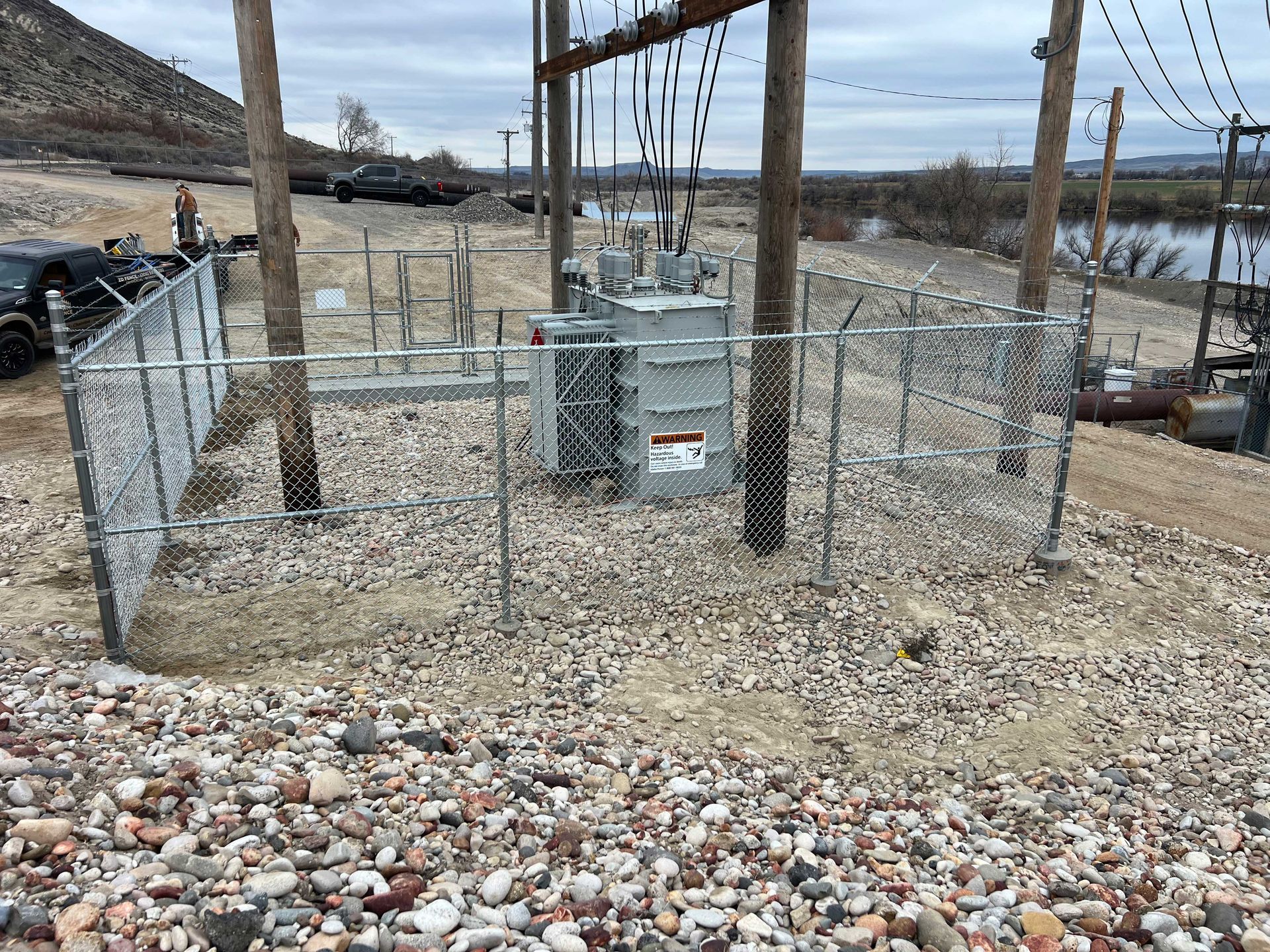Electrical transformer station enclosed by a chain-link fence on a gravel surface. Power lines overhead, overcast sky.
