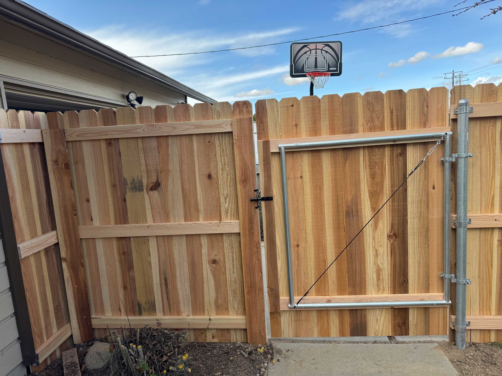 Wooden fence with two gates, one metal-framed, with a basketball hoop visible in the background against a blue sky.