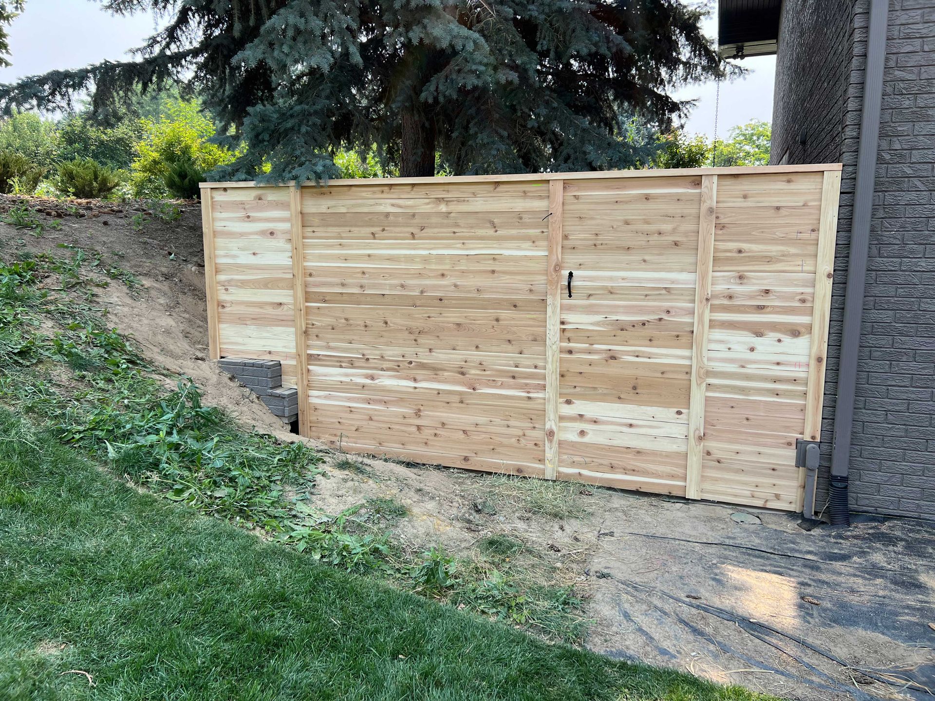 Wooden privacy fence next to a building, partially blocking a sloping yard.