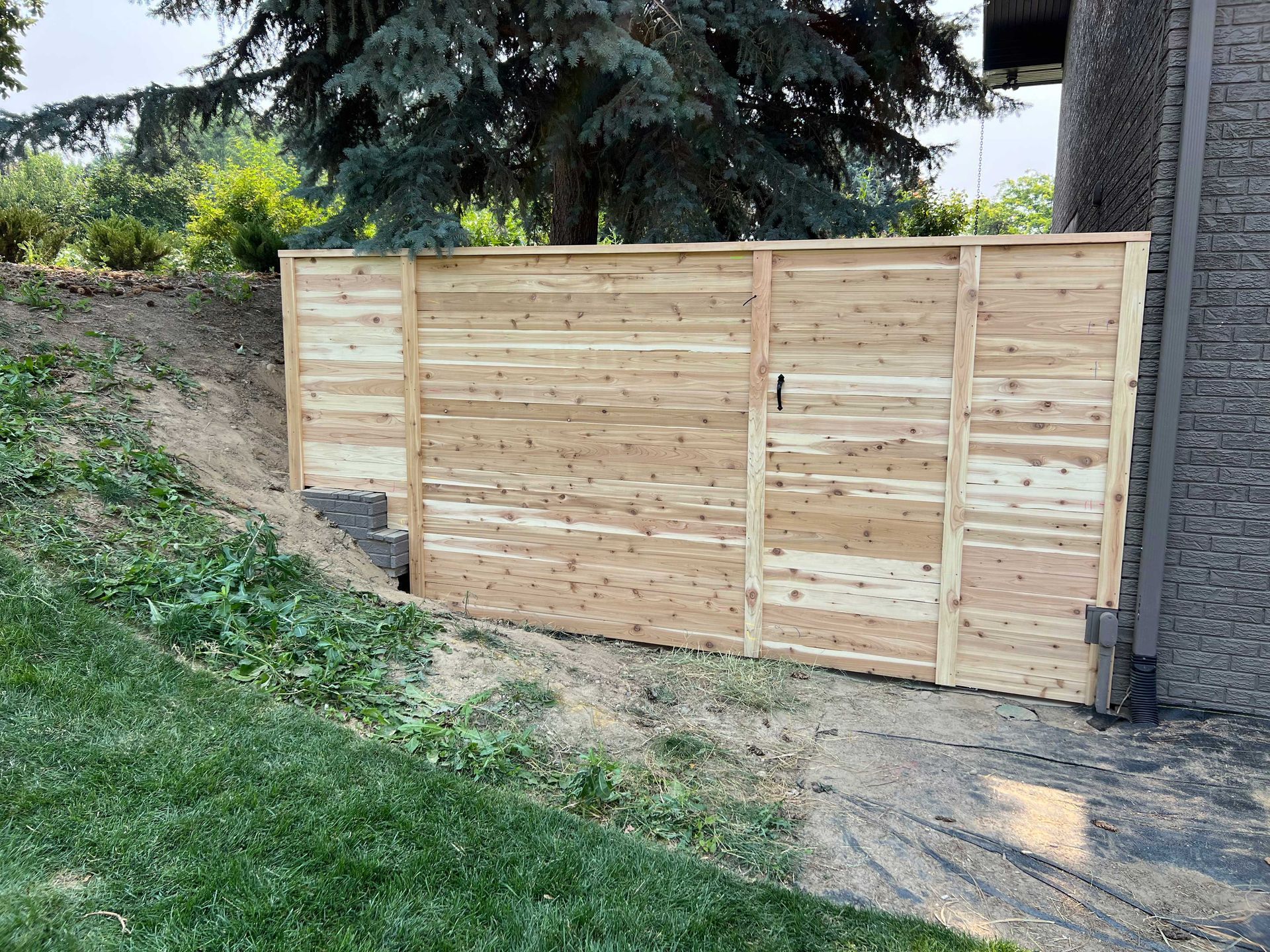 Wooden fence with a gate beside a building and a grassy hill.