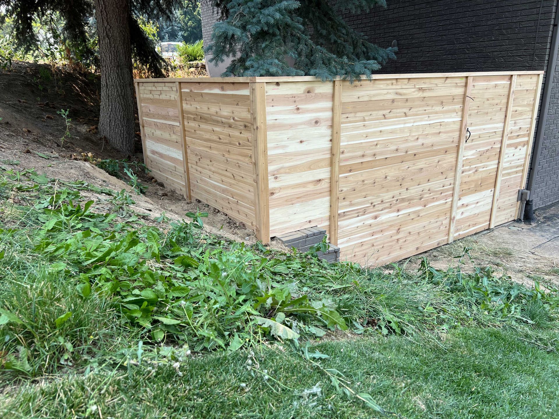 Wooden fence on a slight hill, next to a tree and building. Green grass and weeds are in front.