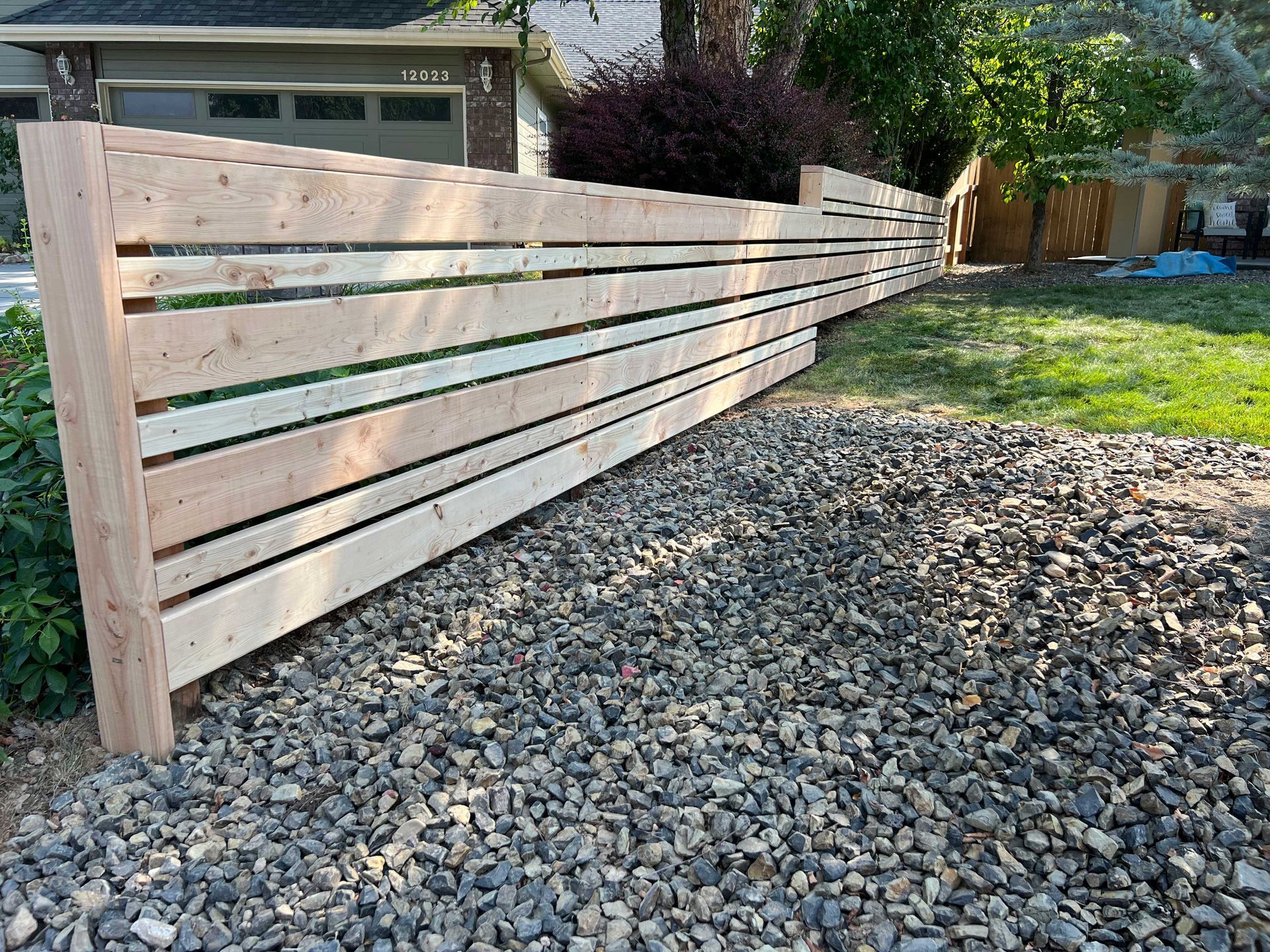Wooden horizontal slat fence in front of a house, gravel ground.