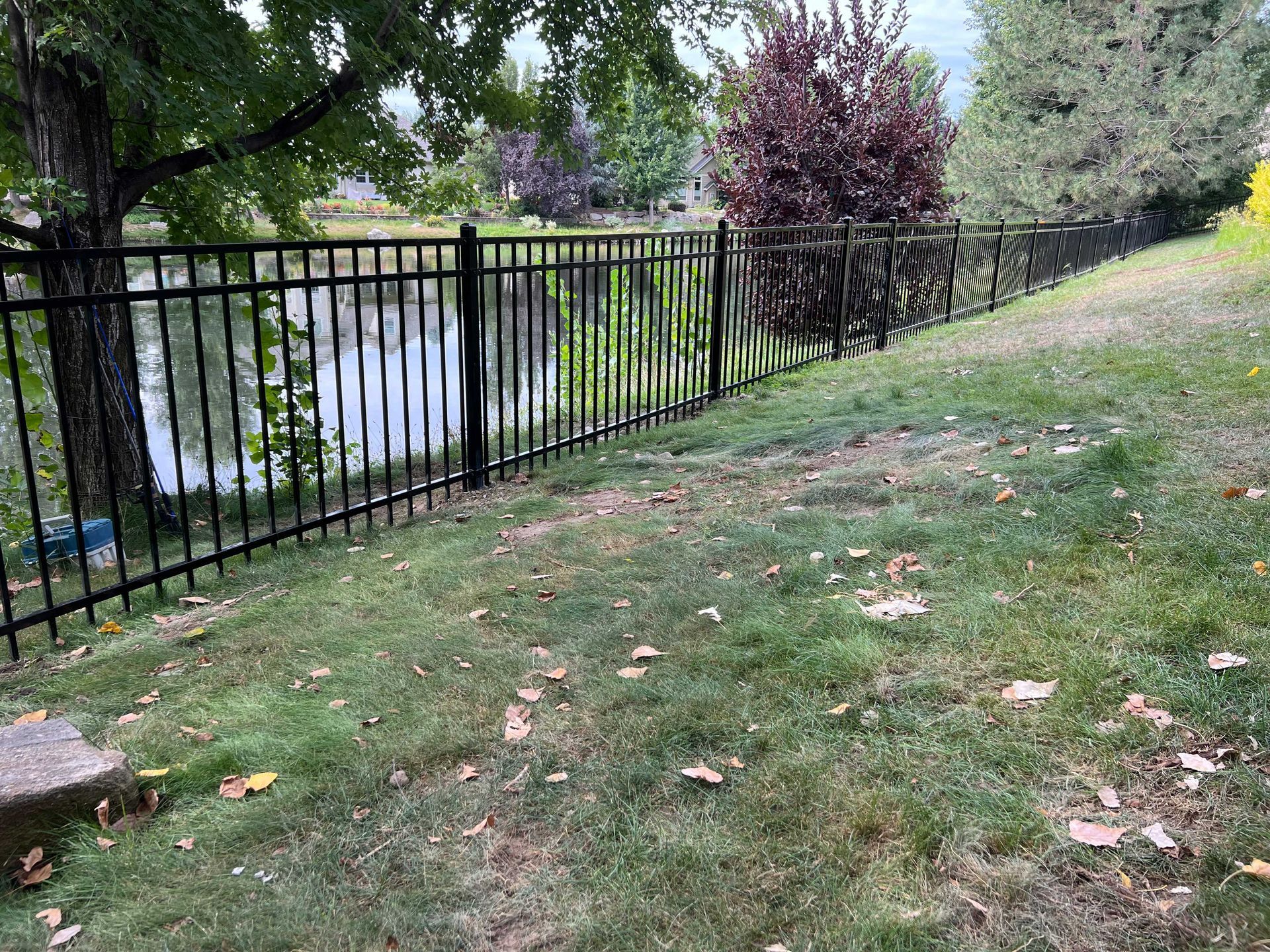 Black metal fence along a grassy slope, overlooking a body of water and trees.