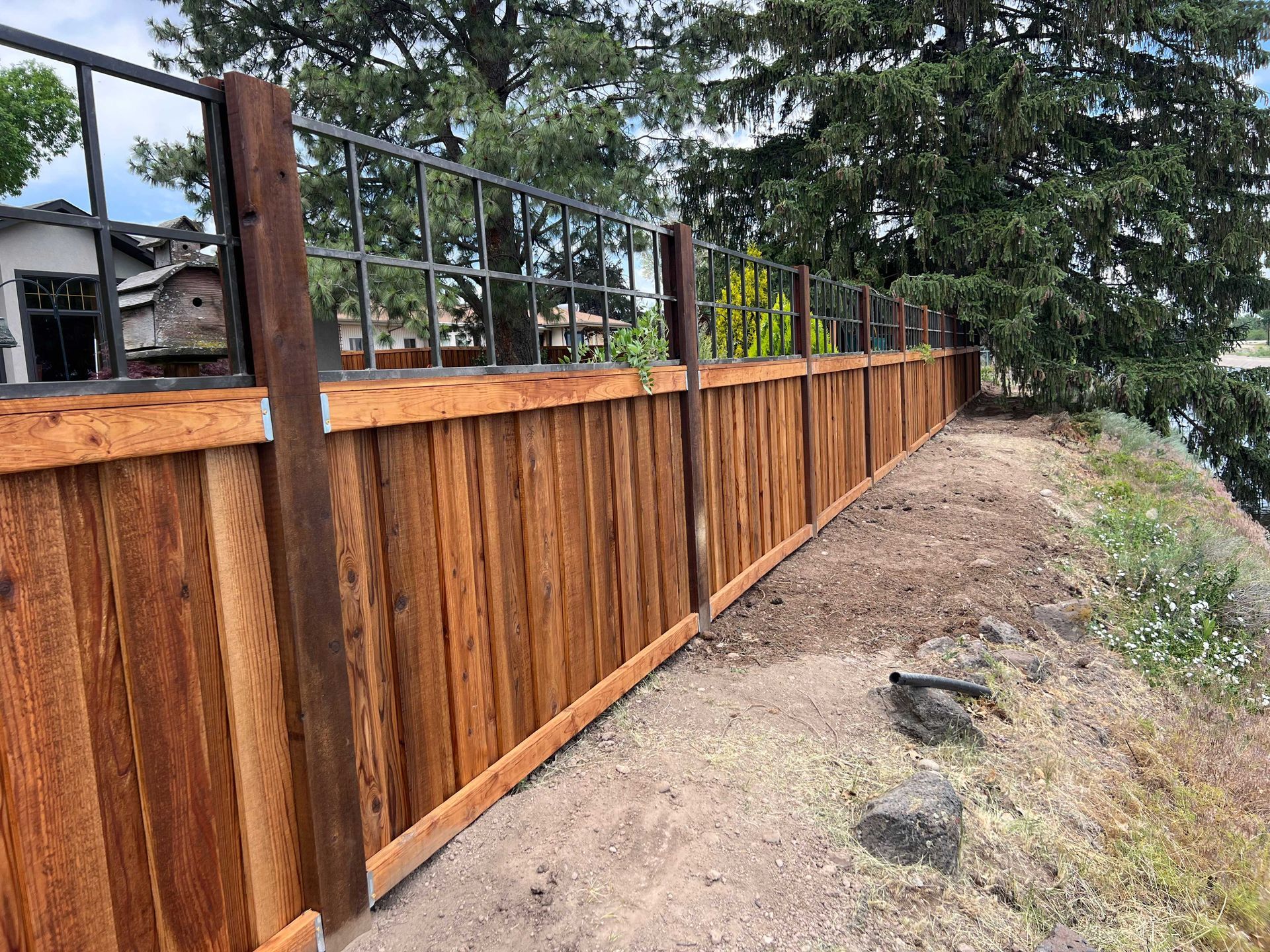 Wooden fence with metal top, along a grassy embankment, near trees and buildings.