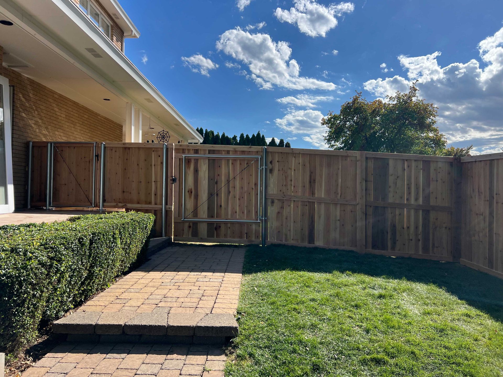 Brick pathway leads to wooden fence with gate in backyard. Green grass, blue sky.