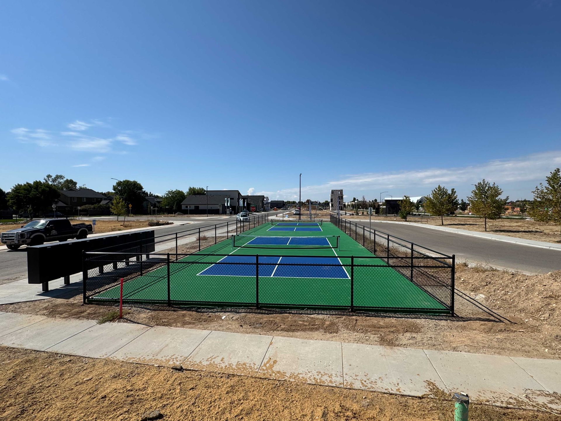 Pickleball courts with green and blue surfaces, surrounded by a black fence, in a sunny, residential area.