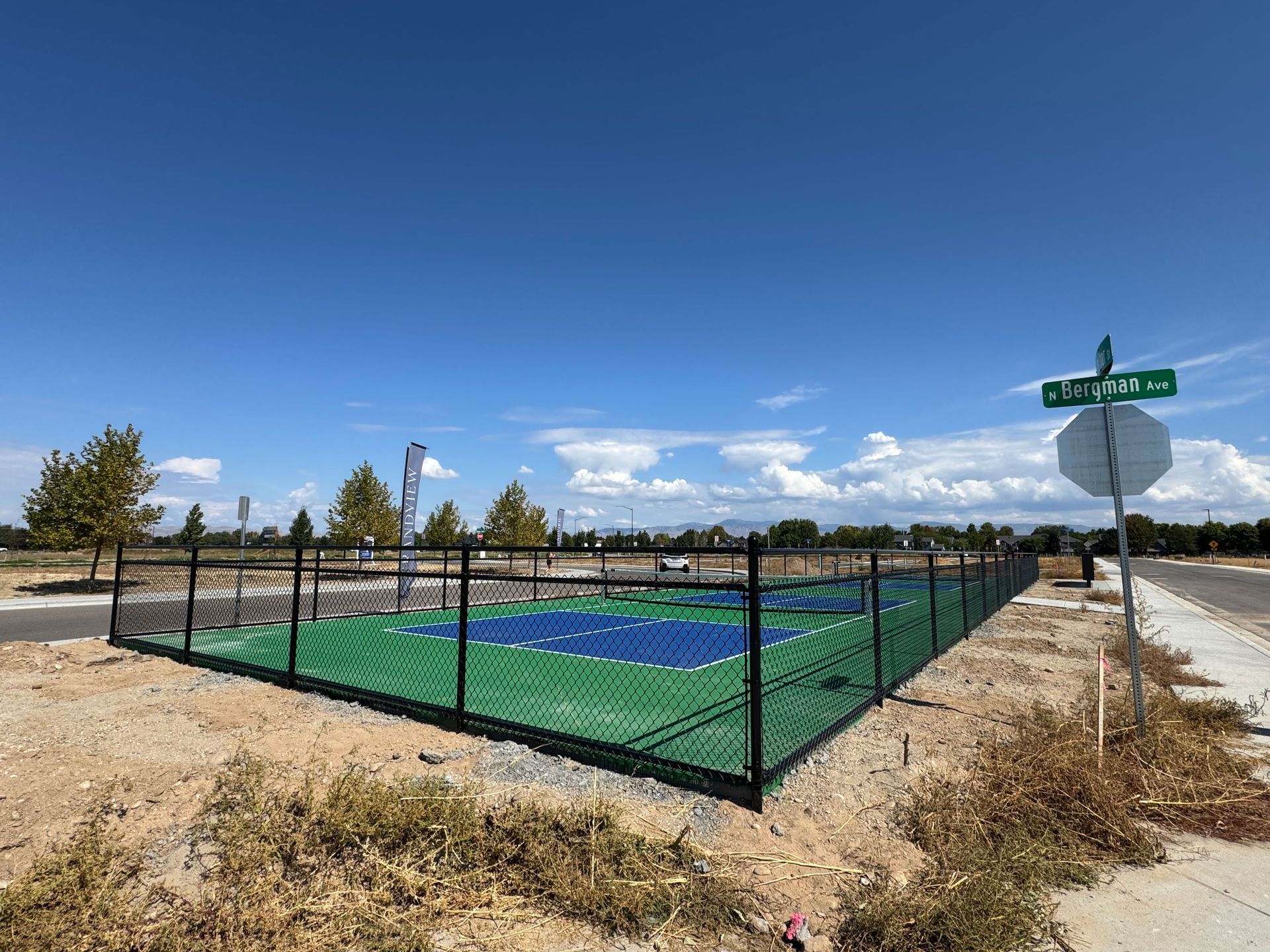 Fenced outdoor basketball court with green and blue playing surface, on a corner lot under a blue sky.