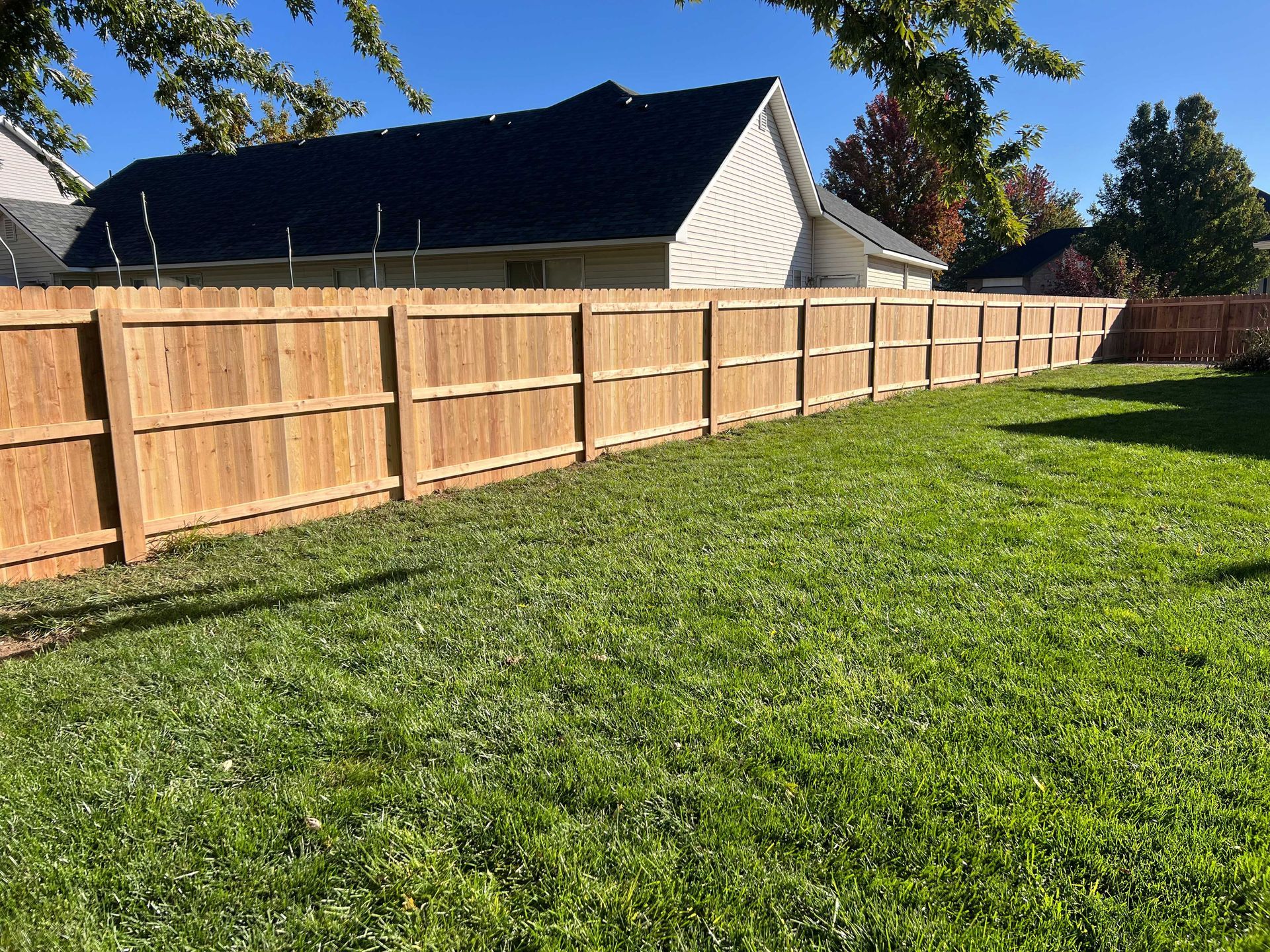 Wooden fence in a grassy yard, with a house and trees in the background under a blue sky.
