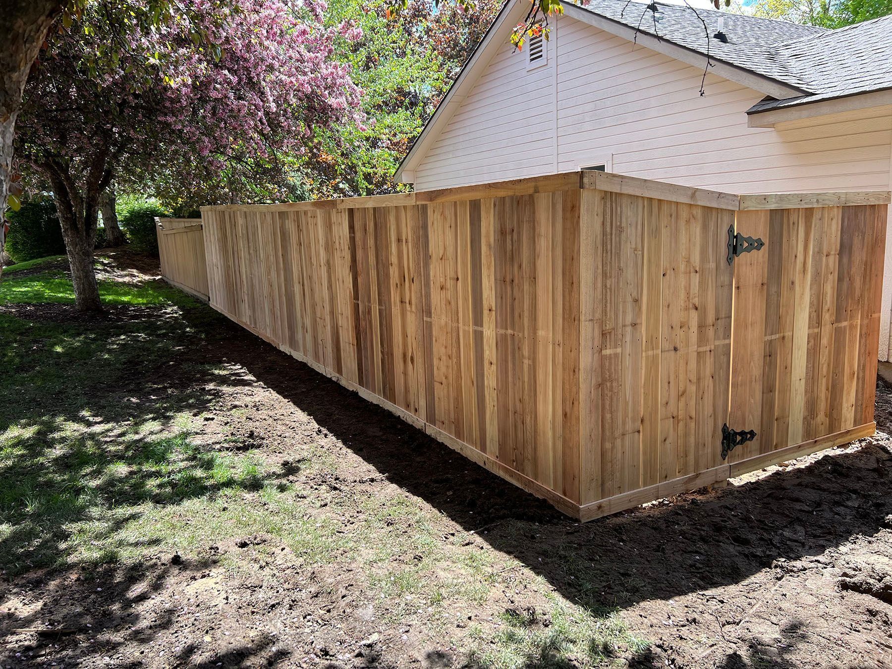 Wooden fence along the side of a white house with a partially visible tree in a grassy yard.