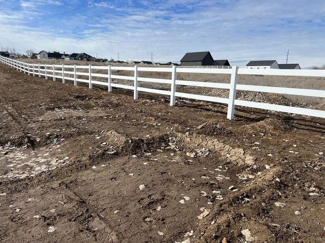 White fence in a field with brown dirt, with buildings and a blue sky in the background.