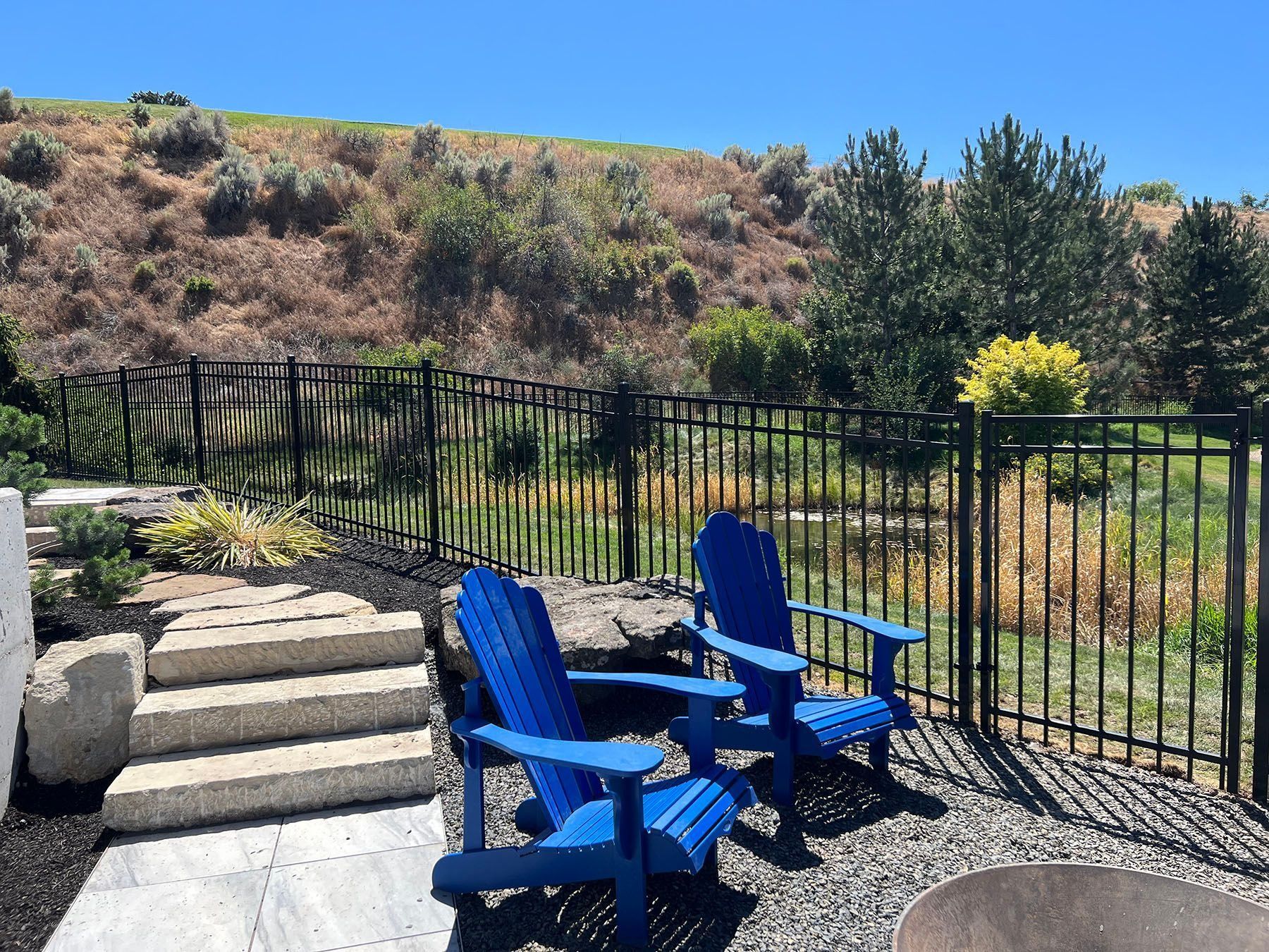 Two blue Adirondack chairs on a patio, overlooking a fenced yard and hillside under a blue sky.