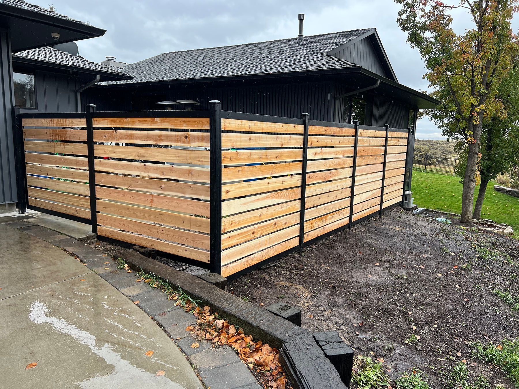 Wooden horizontal slat fence with black posts, next to a house with black siding.
