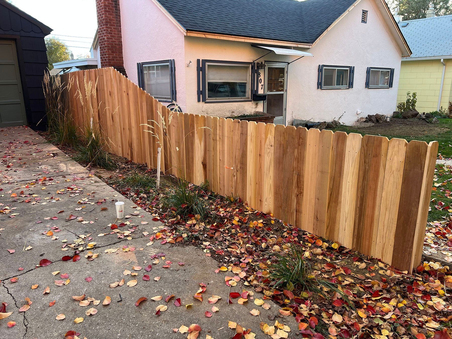 Wooden fence along the side of a beige house with a dark roof; leaves on the ground.