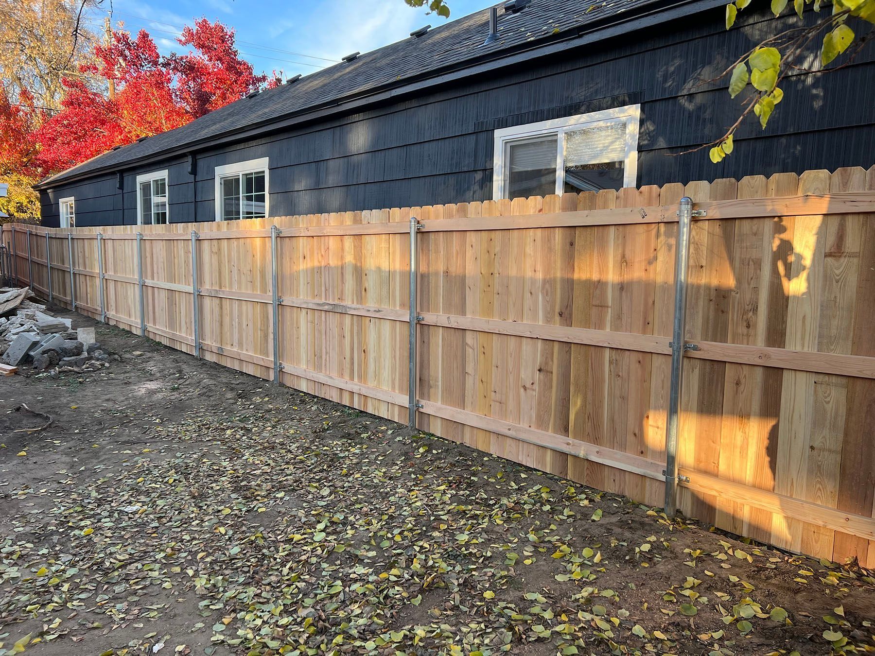 Wooden fence along a dark-painted building with colorful fall foliage in the background.