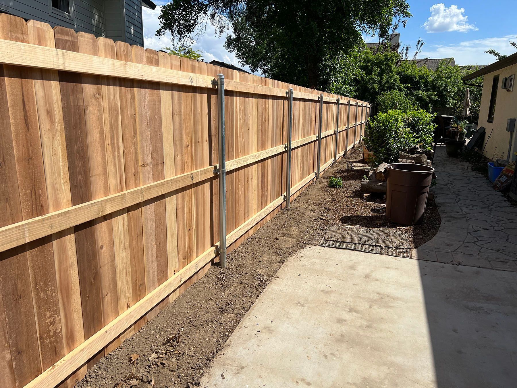 Wooden fence along a concrete path, with dirt and small plants in between. Blue sky in background.