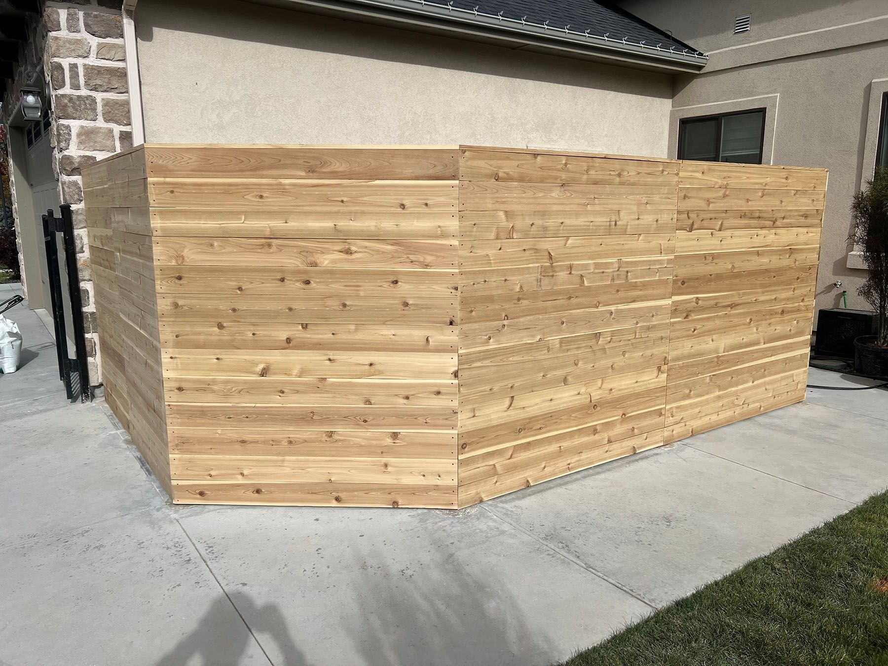 Wooden privacy fence against a beige wall and building corner, on concrete with grass.