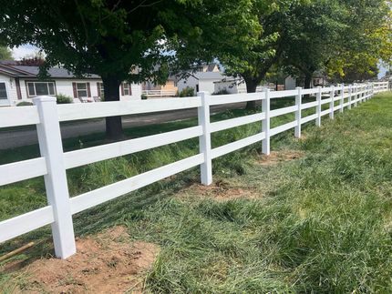 White three-rail fence alongside a grassy area with houses and trees in the background.