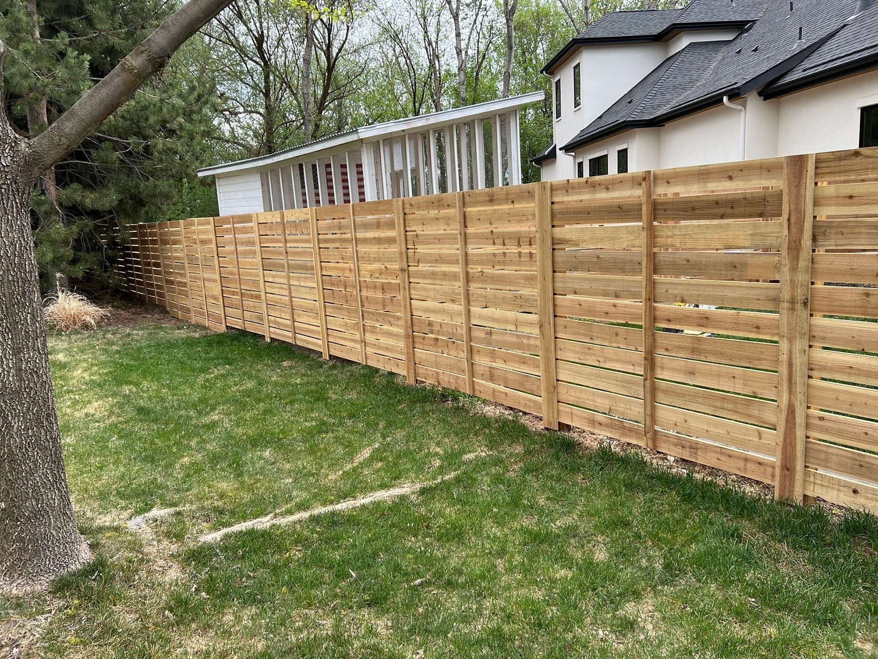 Wooden horizontal slat fence in a grassy yard, beside a house with a dark roof.
