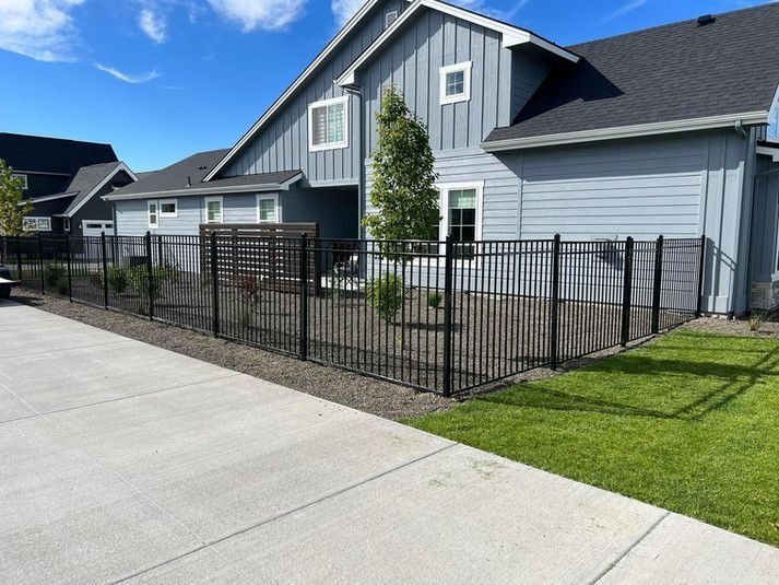 Black metal fence surrounds a house with light blue siding and green lawn on a sunny day.