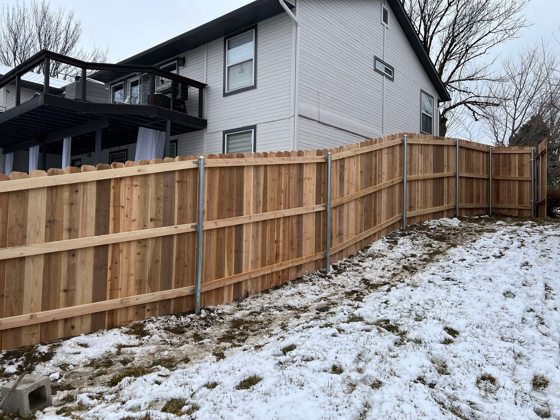 Wooden fence in front of a white house with a black deck. Snow on the ground.
