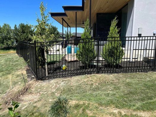 Black metal fence around a yard with trees, plants, and a house in the background.
