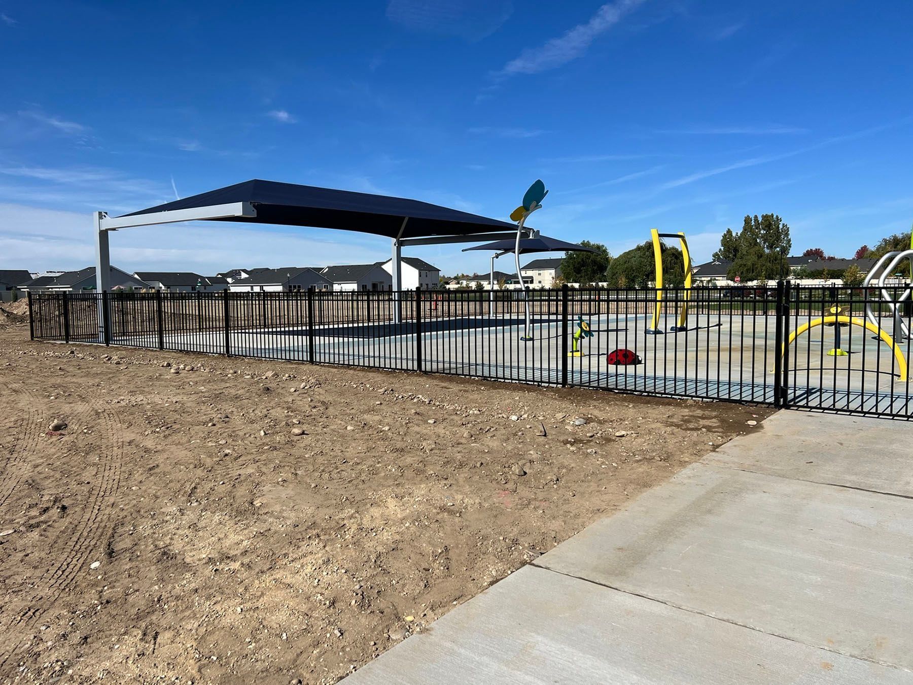 Black fence surrounds a playground with a shade structure and water features on a bright, sunny day.