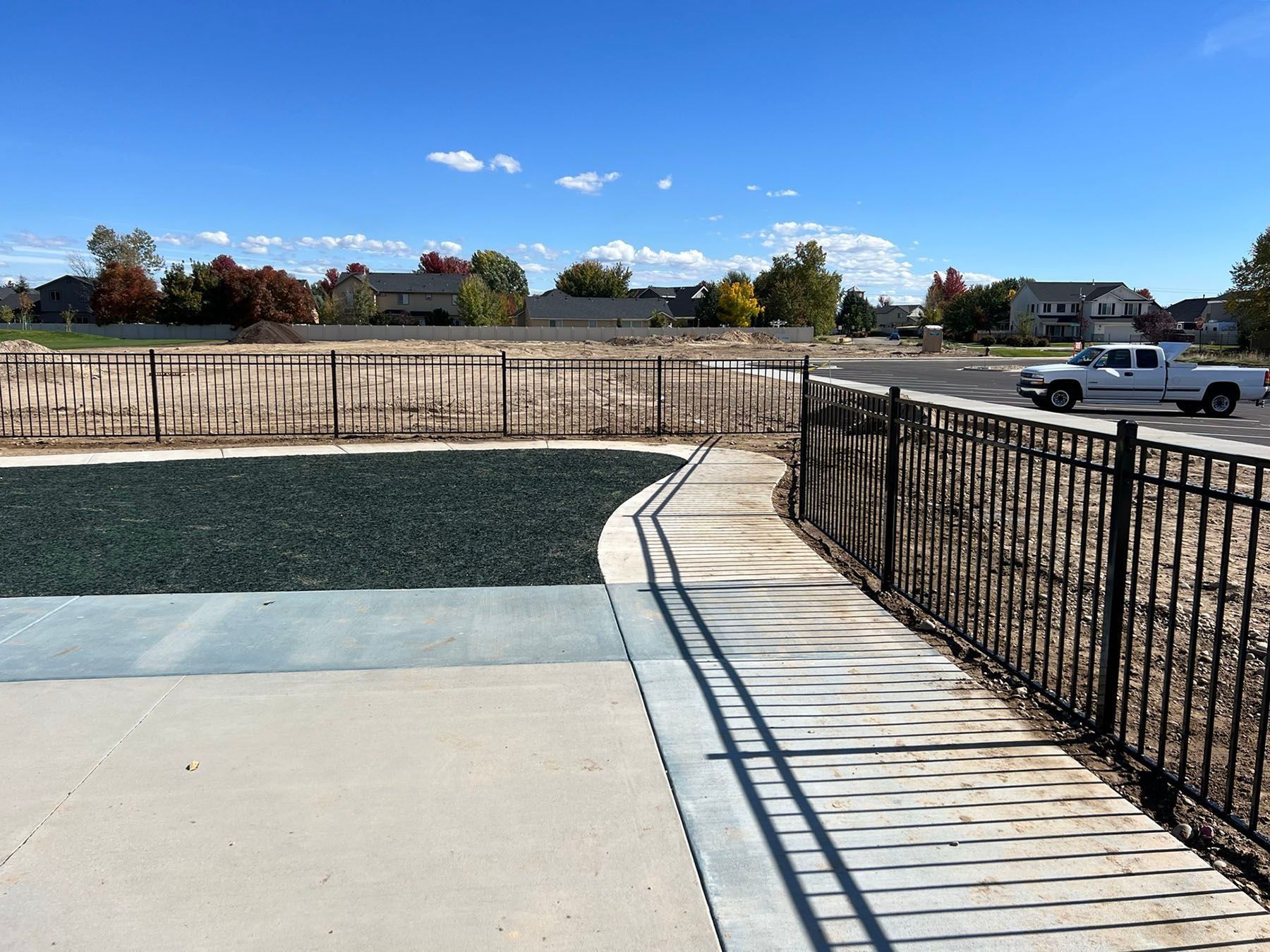 Black metal fence with walkway, playground area with green surface, and construction site. Blue sky.