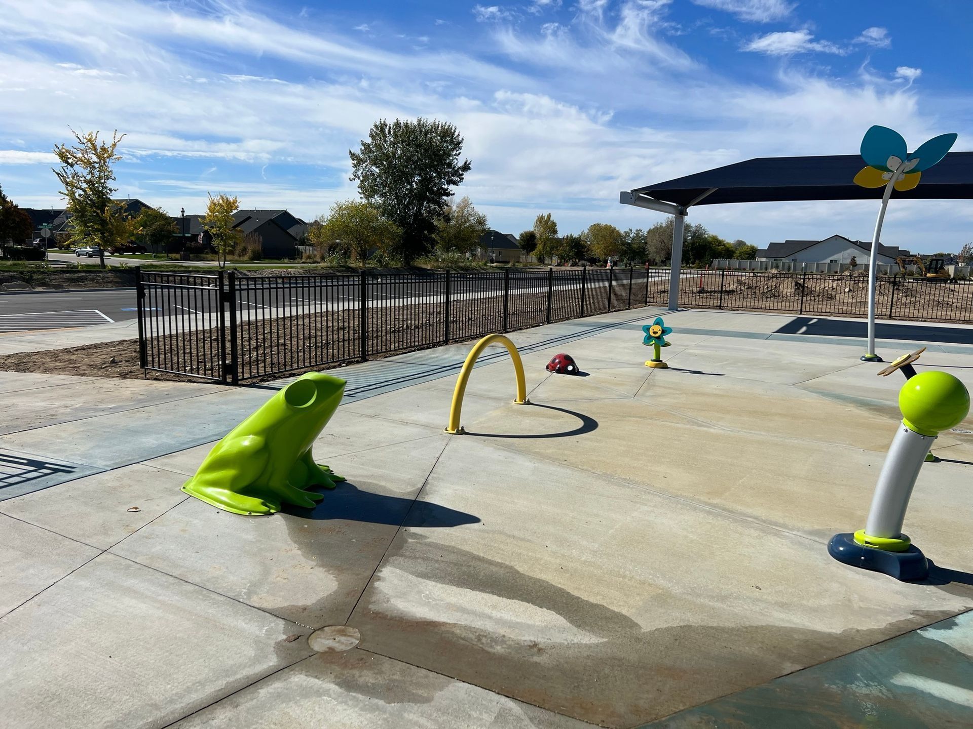 A splash pad at a park with a frog, water features, and a shaded area, under a blue sky.
