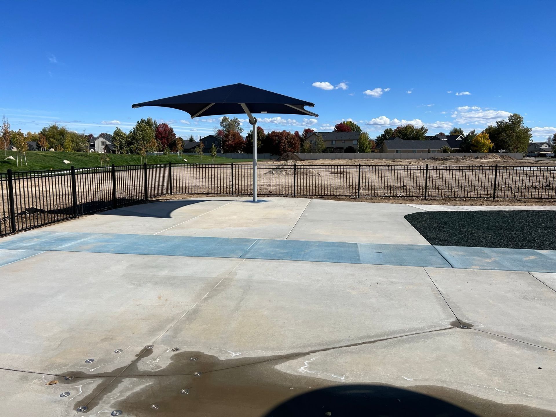 Concrete patio with a dark umbrella on a sunny day; fence and houses in the background.