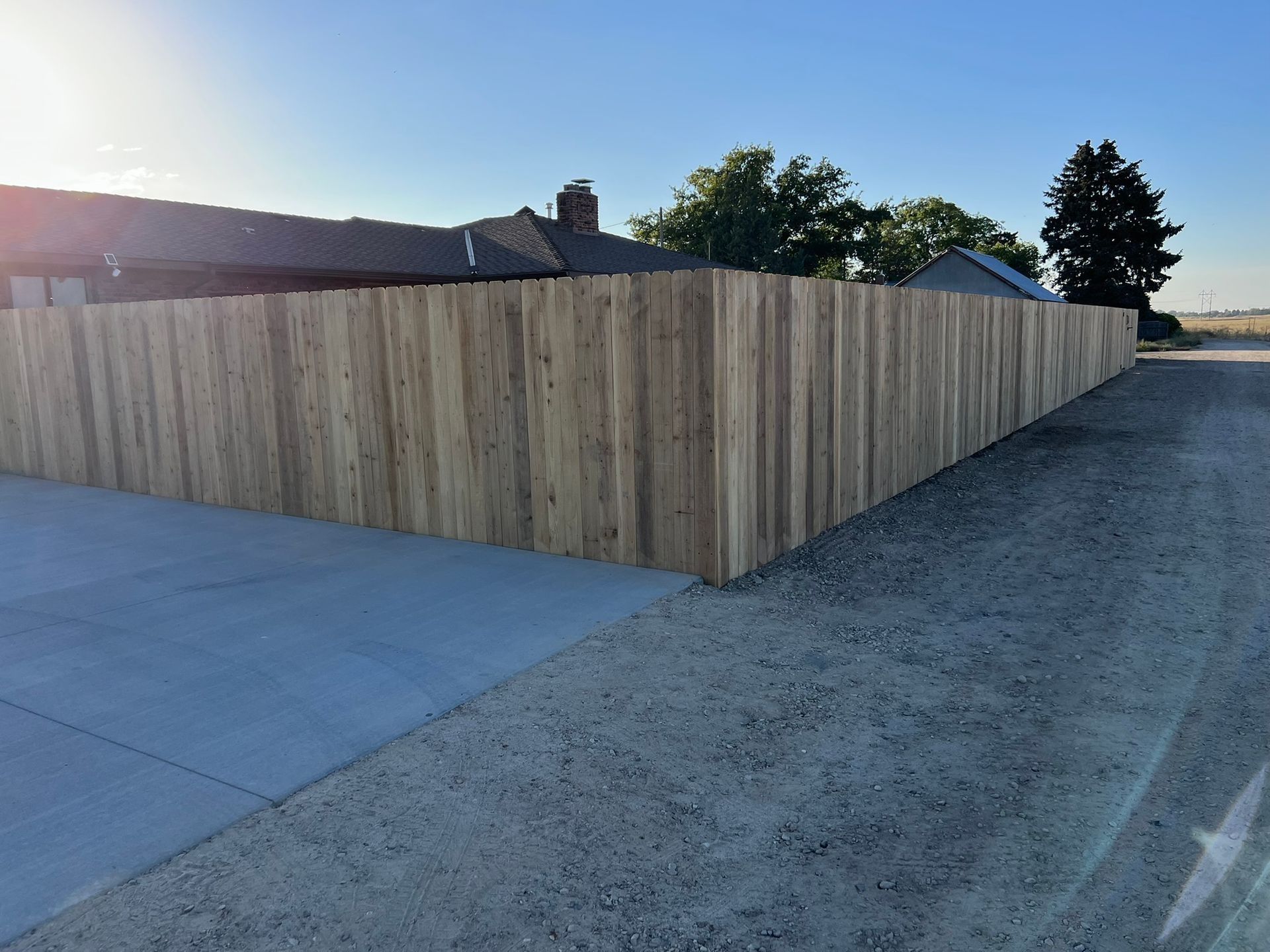 Wooden privacy fence alongside a concrete driveway and gravel road. Sunlight highlights the wood.